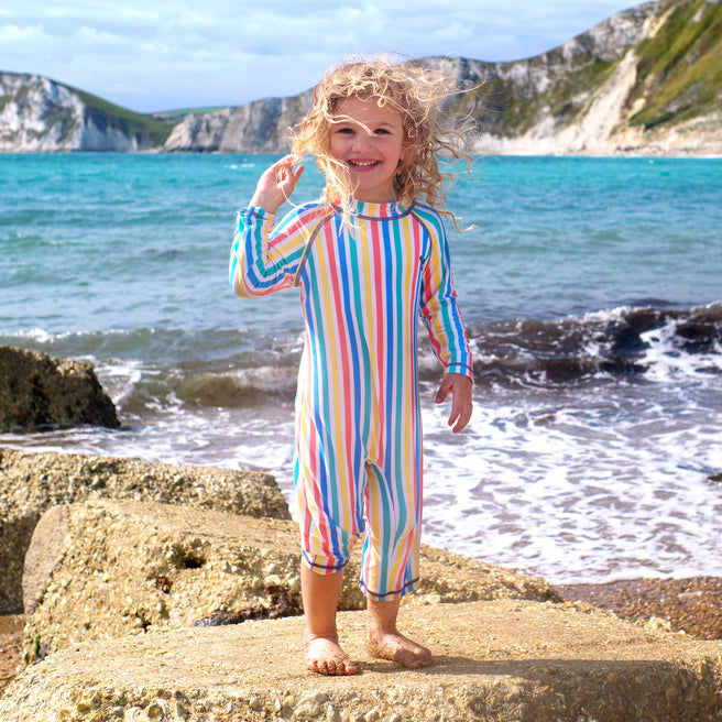 A child wearing a UV Protective Surf Suit Multi Stripe stands on rocks at the beach with the sea and cliffs in the background.